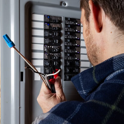 A close-up shot of a technician or handyman using wire strippers to prepare electrical cables for installation in a home circuit breaker box. Concept of home maintenance, electrical safety, and professional trade services.