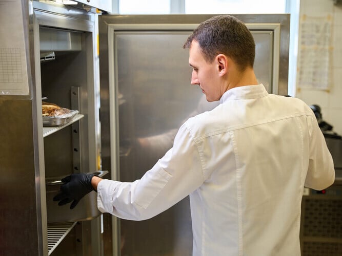 Cook wearing protective gloves puts container of food in refrigerator